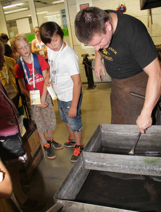 Ein Schmied kühlt ein Metallstück in einem Wasserbecken, während Kinder zusehen.