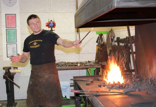 Ein Schmied mit Lederschürze hantiert mit glühendem Metall an einer Esse.