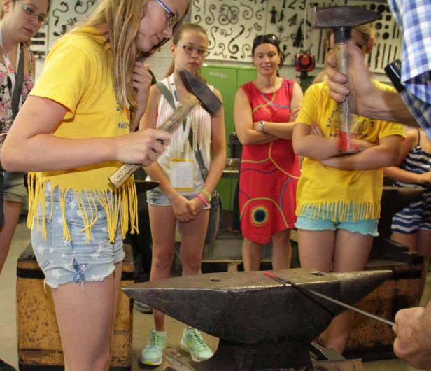 Eine Gruppe von Mädchen steht um einen Amboss und beobachtet das Schmieden.