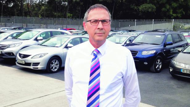 Ein Mann mit Brille und Krawatte steht vor einer Reihe geparkter Autos.