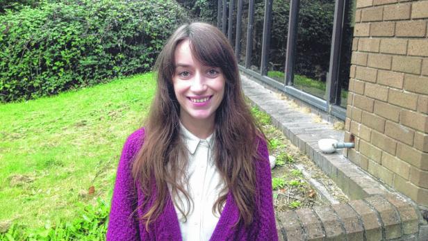 Eine lächelnde Frau mit braunen Haaren und einer lila Strickjacke steht im Freien.