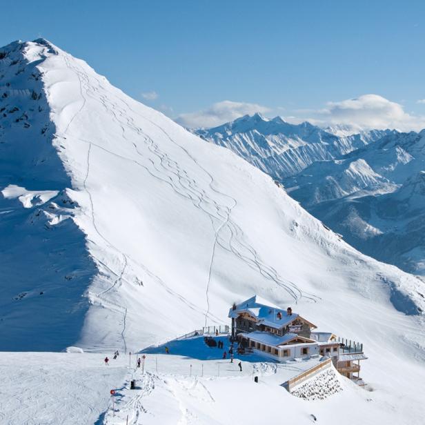 Ein Berghaus liegt unterhalb eines schneebedeckten Berges mit Skifahrern.