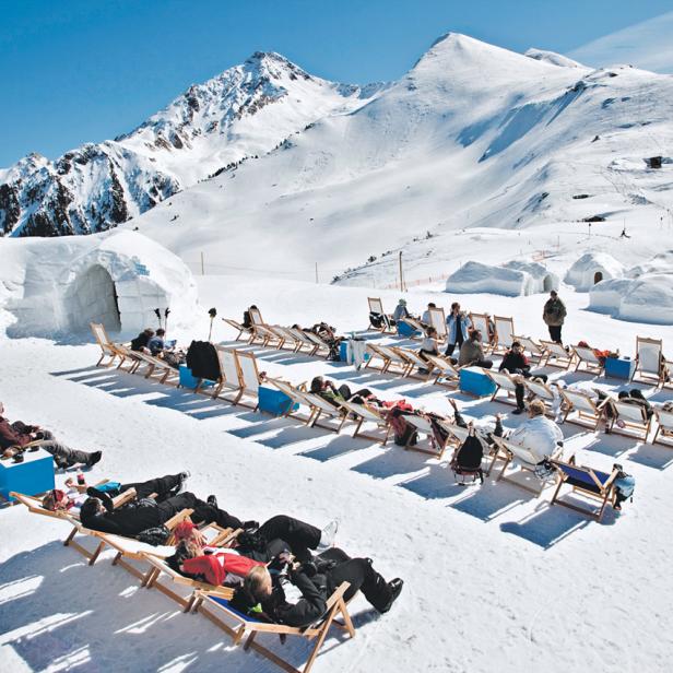 Viele Menschen entspannen sich in Liegestühlen im Schnee vor einer Bergkulisse und Iglus.