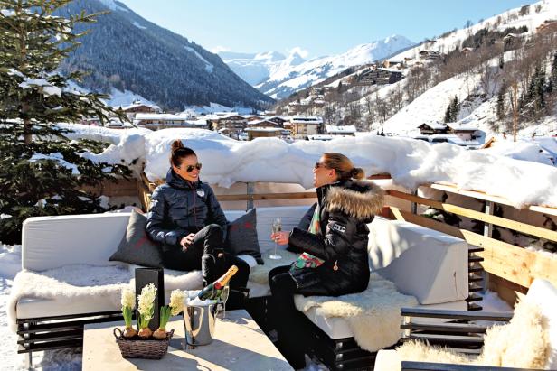 Zwei Frauen sitzen auf einer Terrasse mit Blick auf eine verschneite Berglandschaft und trinken Champagner.