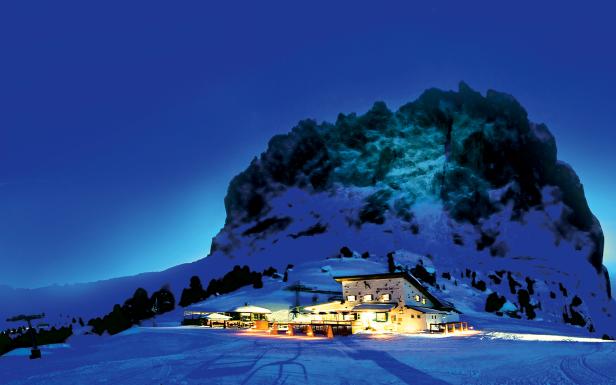 Eine beleuchtete Berghütte unterhalb eines schneebedeckten Berges in der Nacht.