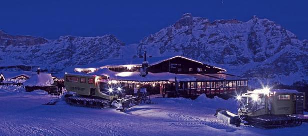 Zwei Pistenraupen stehen vor einem beleuchteten Restaurant in den verschneiten Bergen.