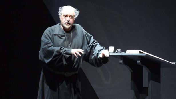 Ein Mann in einem schwarzen Gewand steht auf einer Bühne neben einem Pult.