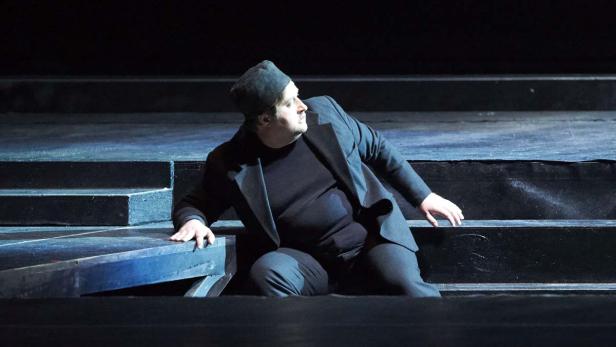 Ein Mann in einem schwarzen Anzug und einer Mütze sitzt auf einer dunklen Treppe.