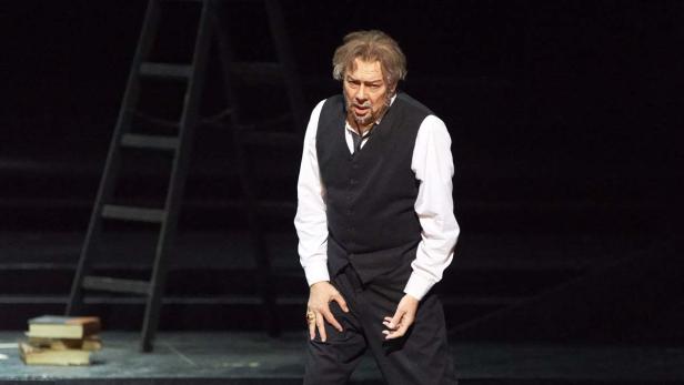 Ein Mann in einem schwarzen Anzug steht auf einer Bühne mit einer Leiter und Büchern im Hintergrund.