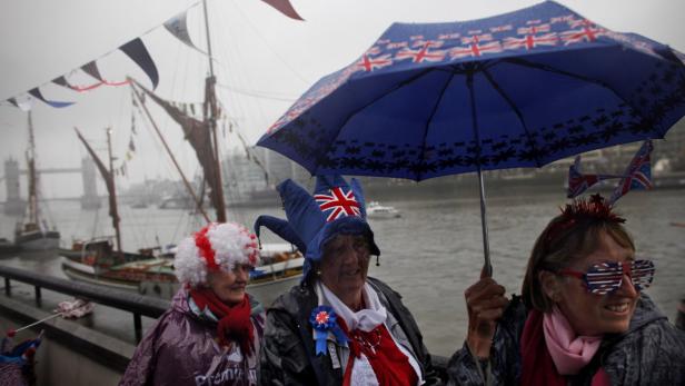 Drei Frauen mit Union-Jack-Dekorationen stehen am Ufer der Themse.