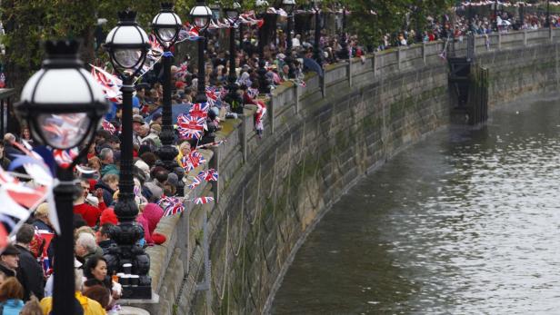 Eine Menschenmenge mit Union-Jack-Flaggen säumt ein steinernes Flussufer.