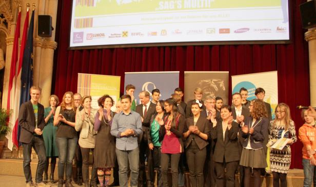 Eine Gruppe von Menschen steht auf einer Bühne und applaudiert bei einer Veranstaltung „Sag’s Multi!“.