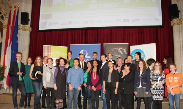 Eine Gruppe von Menschen steht auf einer Bühne vor einem roten Vorhang und einem Bildschirm mit der Aufschrift „SAG'S MULTI!“.