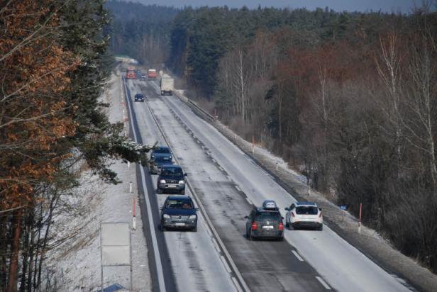 Autos und LKW fahren auf einer verschneiten Straße durch eine Waldlandschaft.
