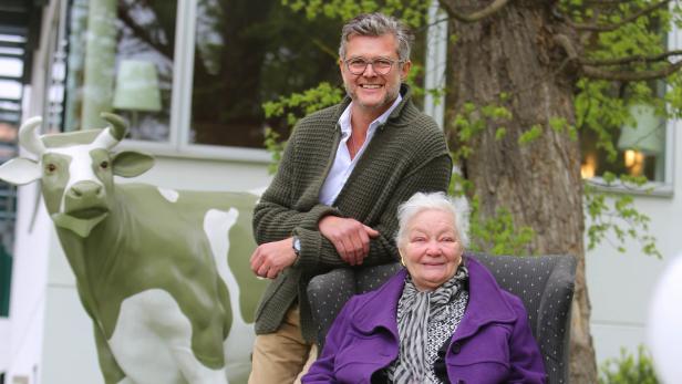 Eine ältere Frau sitzt in einem Sessel, während ein Mann hinter ihr steht; im Hintergrund eine Kuhstatue.