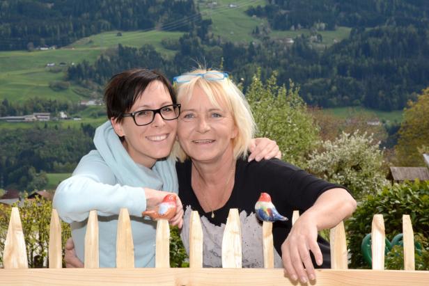 Zwei Frauen umarmen sich vor einem Holzzaun mit Vogeldekoration und einer grünen Landschaft im Hintergrund.