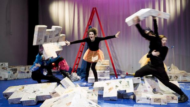 Eine Theaterszene mit mehreren Darstellern und Requisiten aus Zeitungspapier auf einer blauen Bühne.