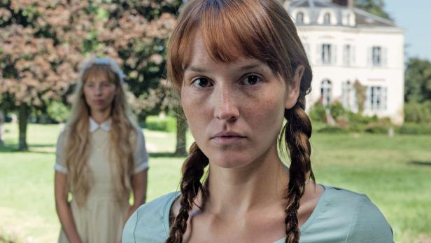Filmseite … Eine junge Frau mit Zöpfen steht vor einem Haus, während eine andere Frau im Hintergrund verschwommen ist.