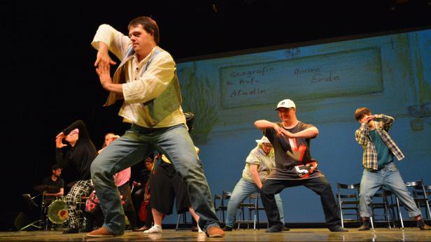 Eine Gruppe von Menschen tanzt auf einer Bühne vor einer Leinwand mit der Aufschrift „Geografie“.