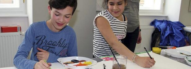 Zwei Kinder malen mit Pinseln und Farben auf Papier.