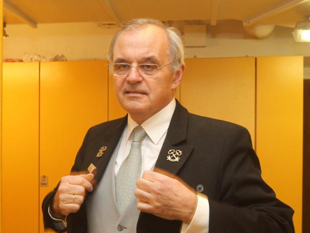 … Ein Mann mit Brille und Anzug mit Abzeichen vor einem gelben Hintergrund.