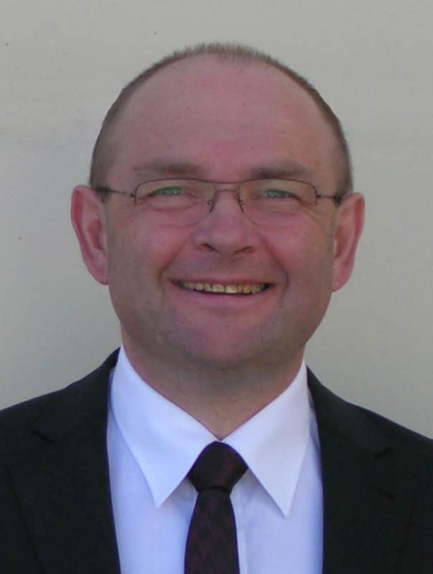 Ein lächelnder Mann mit Brille, Anzug und Krawatte vor einem hellen Hintergrund.