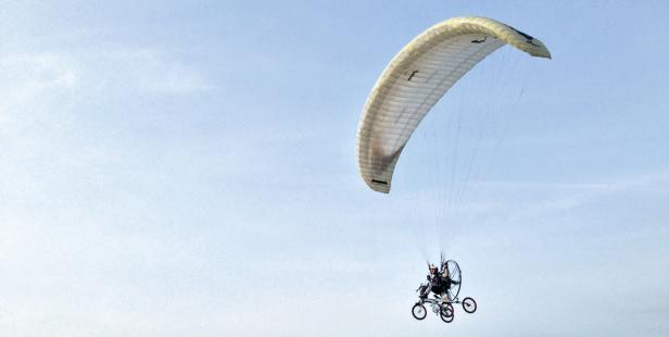 Ein Motorgleitschirm fliegt vor einem hellblauen Himmel.