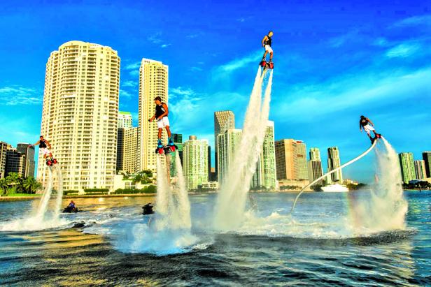 Mehrere Personen betreiben Flyboarding vor der Skyline einer Stadt.