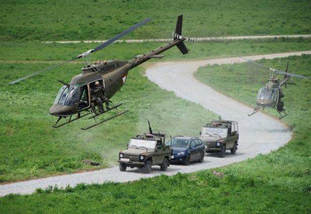 Zwei Hubschrauber begleiten eine Kolonne von Militärfahrzeugen auf einer Schotterstraße.