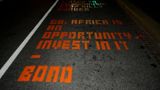 Bono: "G8: Afrika ist eine Chance, investiert darin". Auf der Straße steht in orangefarbener Schrift: „GB: Africa is an opportunity. Invest in it. Bond“.