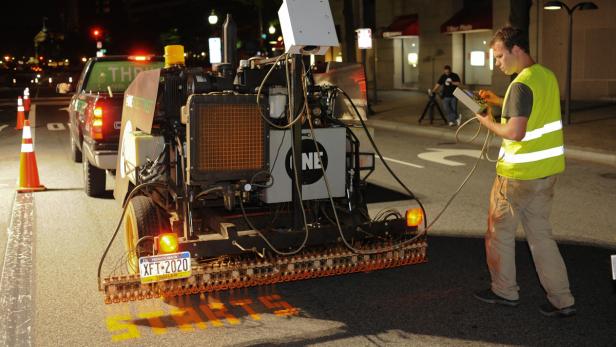 Der Roboter "Street Tweeter" in Aktion. Ein Arbeiter bedient eine Maschine, die gelbe Markierungen auf eine Straße aufbringt.