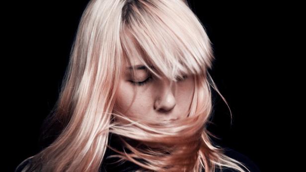 Eine Frau mit blonden Haaren und geschlossenen Augen vor schwarzem Hintergrund.