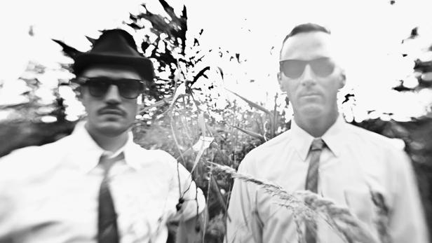 Zwei Männer in Hemden und Krawatten, einer mit Hut und Sonnenbrille, stehen im Freien.