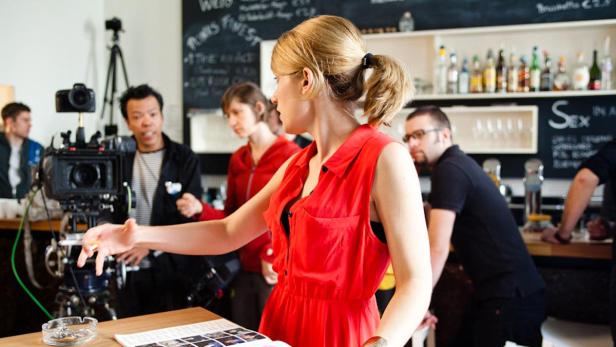 Eine Frau in einem roten Kleid steht vor einer Filmkamera in einem Café.