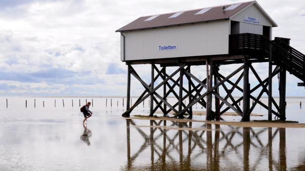 Ein Mann watet im Wasser vor einem Pfahlbau mit der Aufschrift „Toiletten“.