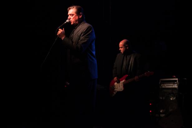 Ein Sänger mit Mikrofon und ein Gitarrist treten auf einer dunklen Bühne auf.