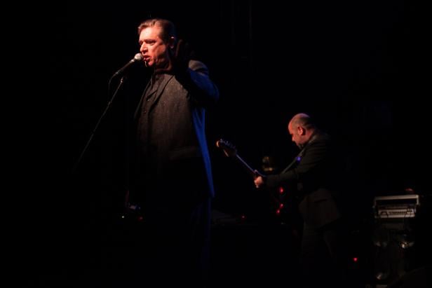 Ein Sänger performt mit einem Gitarristen auf einer dunklen Bühne.
