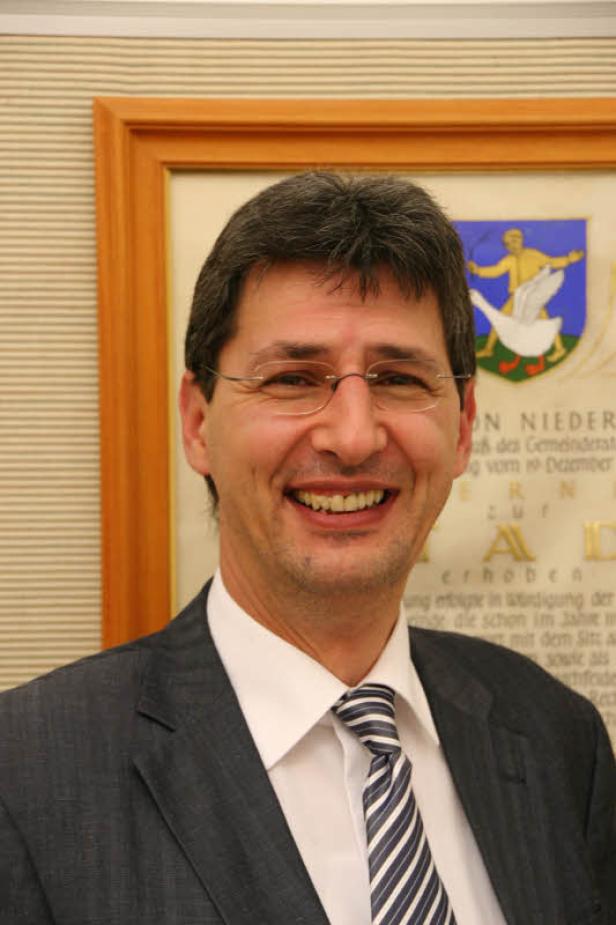 Ein lächelnder Mann mit Brille und Anzug vor einem Wappen.