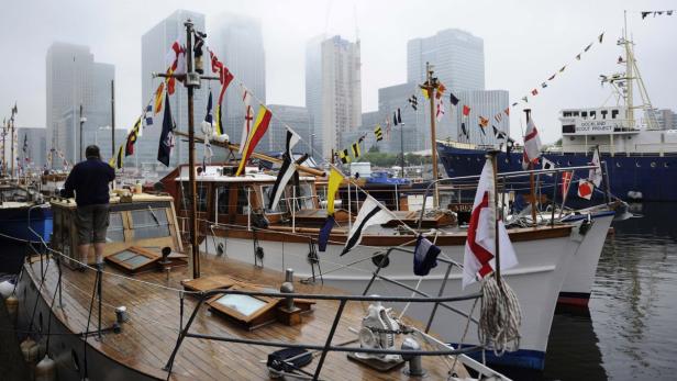Mehrere mit Flaggen geschmückte Boote liegen im Hafen vor der Skyline von London.