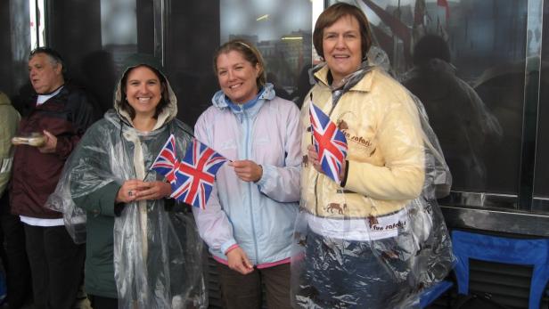 Drei Frauen mit Union-Jack-Flaggen stehen draußen und tragen Regenkleidung.