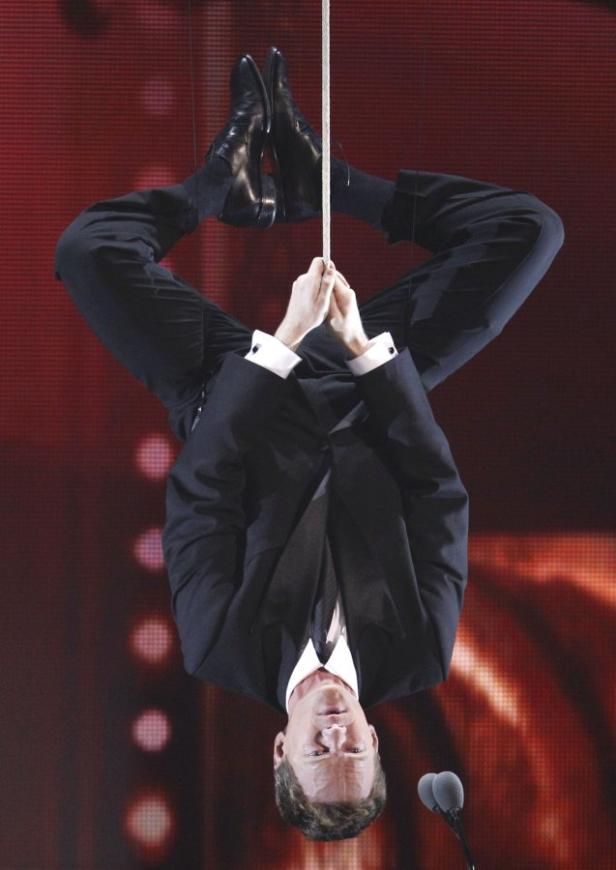 Ein Mann im Anzug hängt kopfüber an einem Seil vor einem roten Hintergrund.