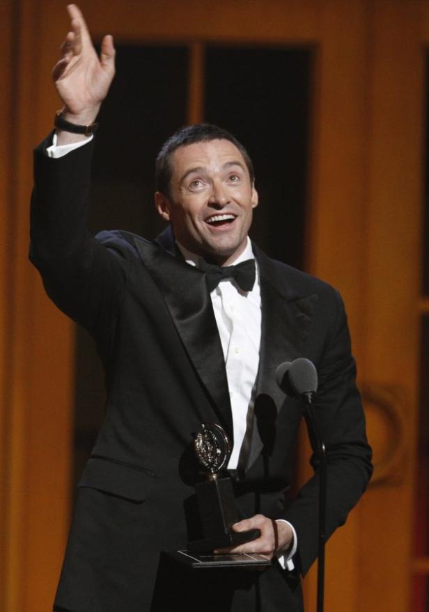 Hugh Jackman hält eine Tony Award Trophäe in der Hand und hebt den Arm.