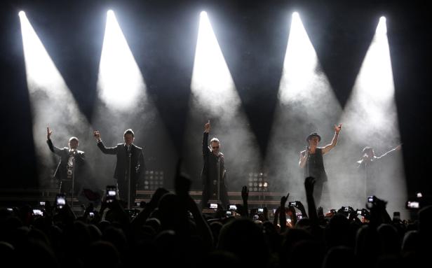 Eine Band tritt vor einem jubelnden Publikum auf der Bühne auf, angestrahlt von Scheinwerfern.