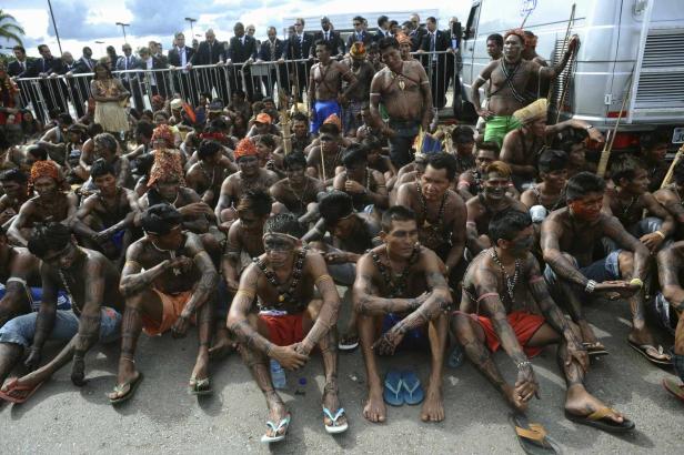 Eine Gruppe indigener Männer sitzt auf dem Boden, viele mit traditioneller Körperbemalung.