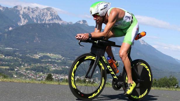 Ein Triathlet fährt mit seinem Rennrad eine Straße entlang, im Hintergrund sind Berge zu sehen.