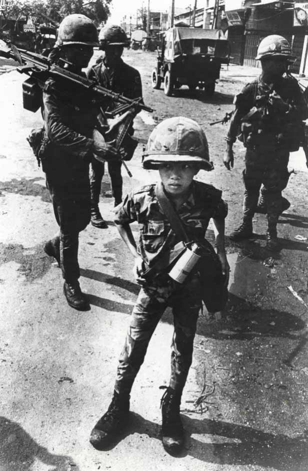 Ein junger Soldat in Tarnkleidung steht inmitten von bewaffneten Soldaten.