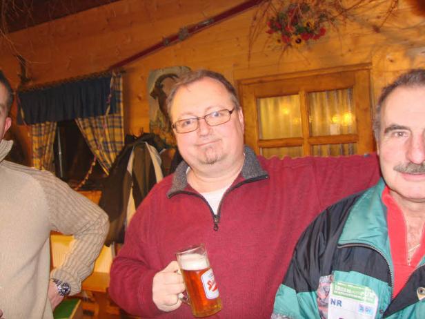 Ein Mann im roten Pullover hält ein Bier in einem holzgetäfelten Raum.