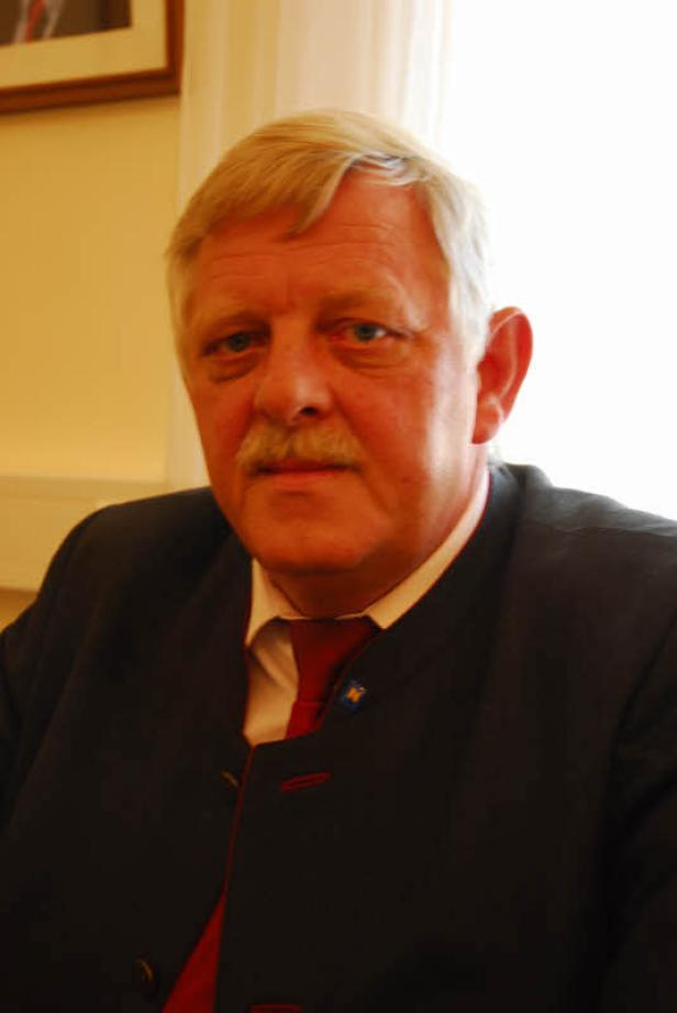 Ein Mann mit Schnurrbart trägt einen dunklen Anzug und eine rote Krawatte.