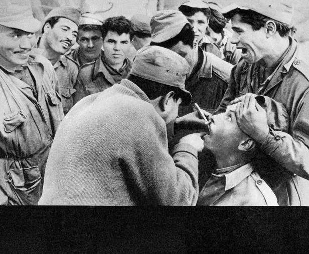 Ein Arzt untersucht den Hals eines jungen Mannes, umgeben von anderen Männern in Uniformen.