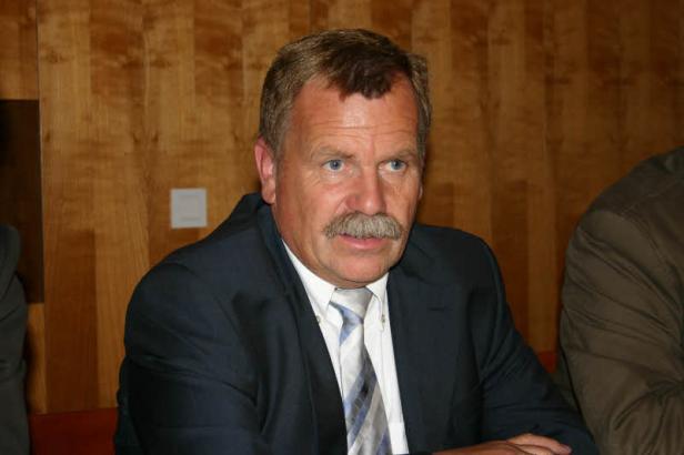 Ein Mann mit Schnurrbart trägt einen Anzug und ein gestreiftes Hemd.